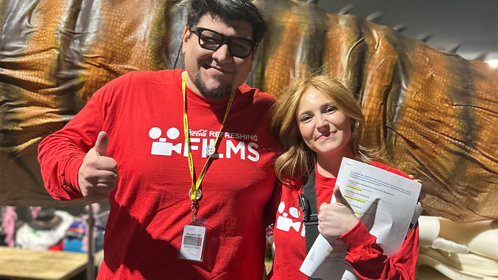 Director Angel and Producer Kylie smile in front of a large animatronic dinosaur tail on set during their film shoot.