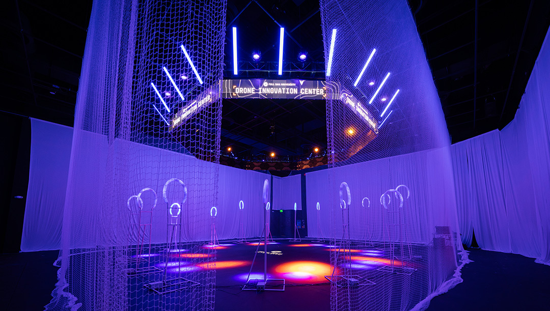 Full Sail’s Drone Innovation Center lit in blue and purple, with safety nets and glowing racing gates arranged inside a large indoor flight zone.