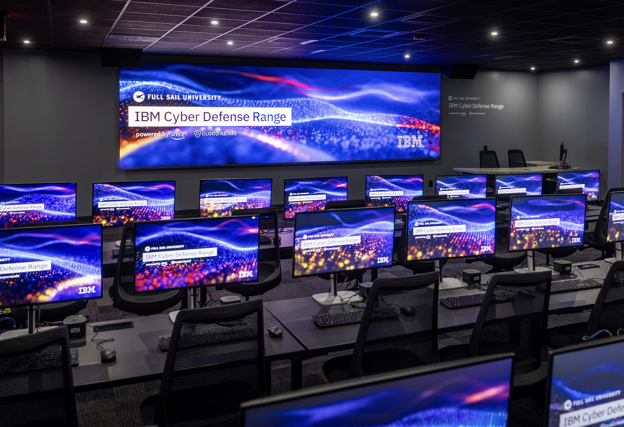 A spacious room with computers and monitors on tables. There is also a large LCD screen on the wall with "Full Sail University IBM Cyber Defense Range Powered by AWS Cloud Range" displayed on the screens.