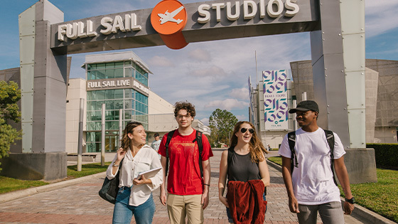 A group of smiling students walks under the Full Sail Studios arch on Full Sail's campus. The Full Sail Live venue is behind the arch.
