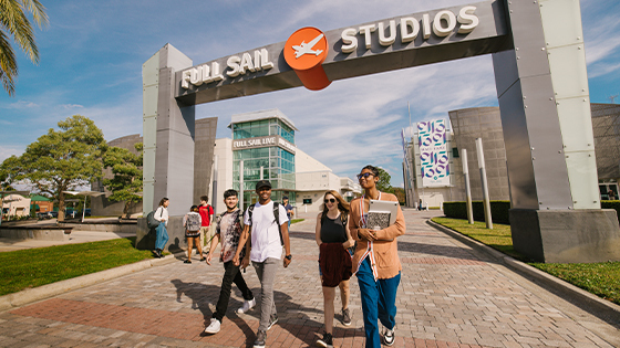 A group of smiling students walks under the Full Sail Studios arch on Full Sail’s campus. The Full Sail Live venue is behind the arch.